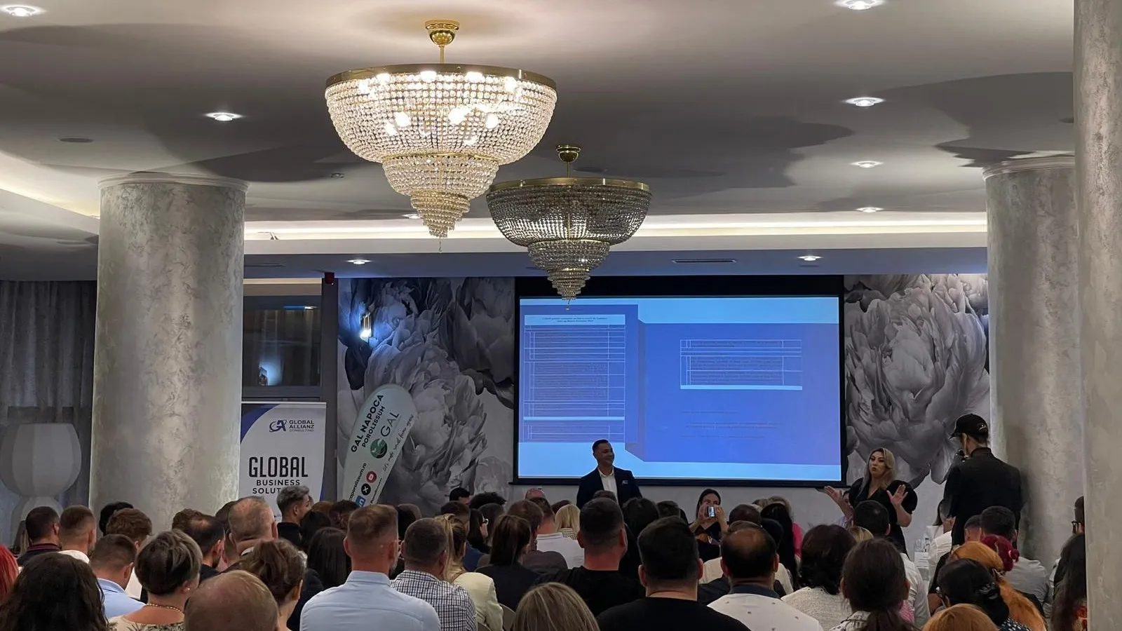 Sală de conferință plină la o sesiune de informare despre finanțări nerambursabile, cu roll-up-ul Global Allianz Consulting.