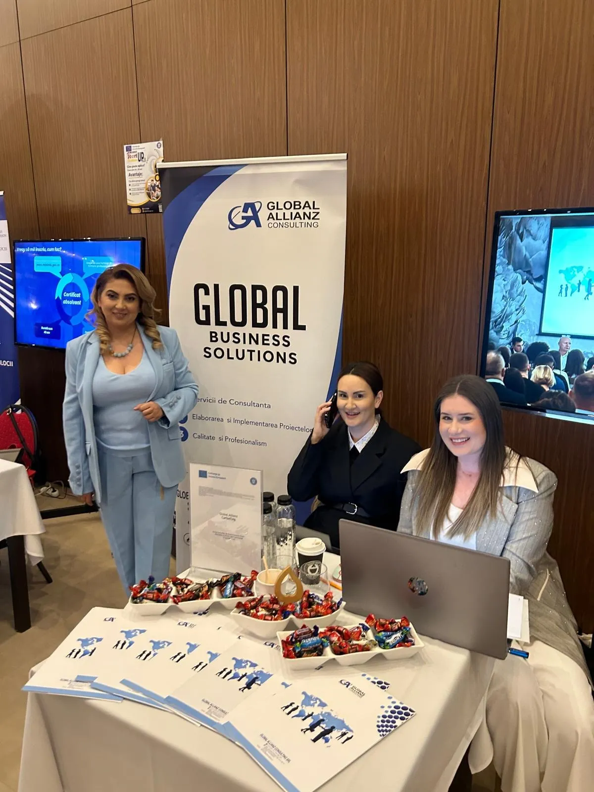 Trei membre ale echipei Global Allianz Consulting la standul firmei, cu broșuri și laptop.
